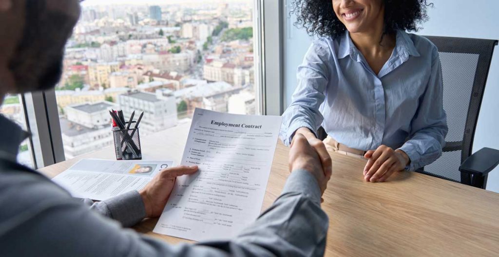 Image of a person shaking hands. They are sitting at a deck. One person is holding an Employment Contract. 