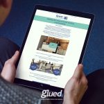 A person sits on a blue carpeted floor, holding a tablet displaying an online brochure titled Making the most of home working environments by Glued Marketing. The screen shows a mix of text, images, and a play button for a video. The word glued is visible at the bottom.