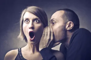 A man whispers into a womans ear, causing her to look surprised with wide eyes and an open mouth—an expression as if shes just heard revealing customer feedback. The background is a textured gray, and both are dressed in dark clothing.