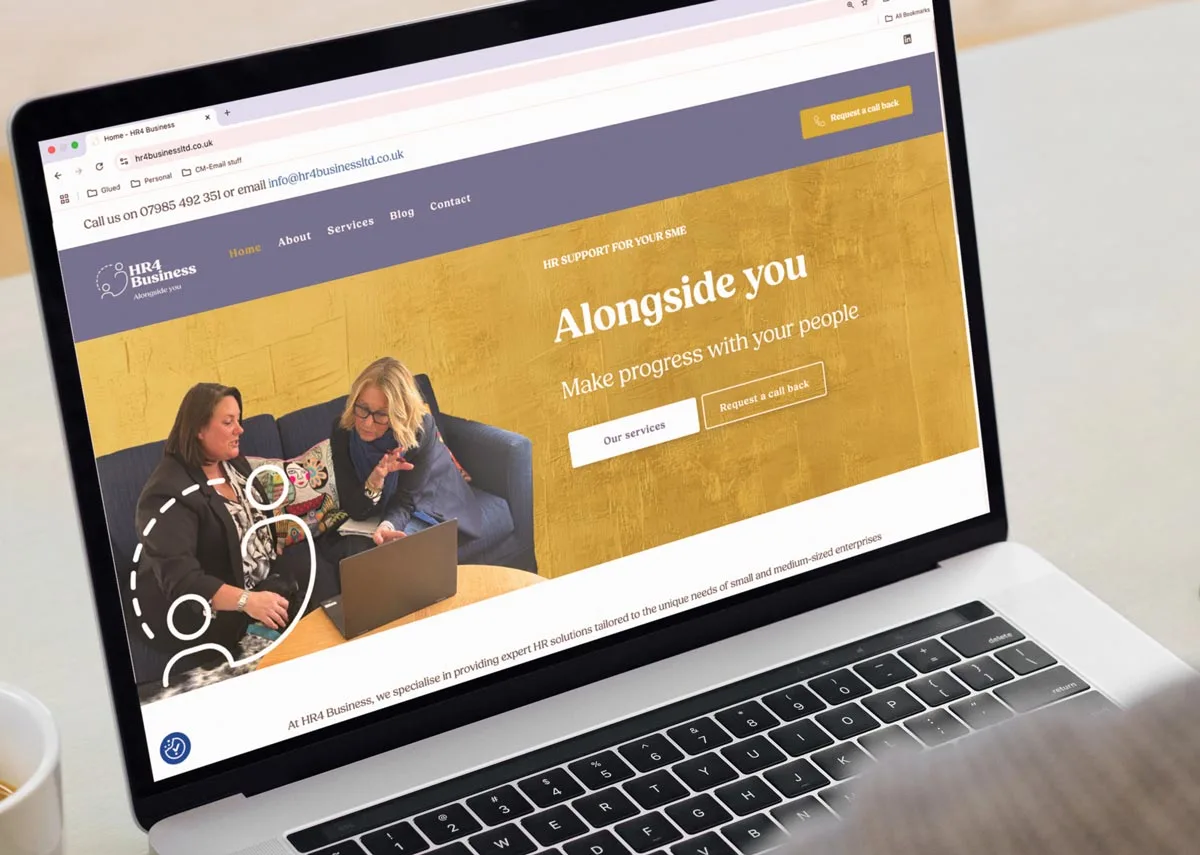 A person uses a laptop displaying a business website with a gold banner that reads “Alongside you: Make progress with your people.” The homepage highlights brand creation for HR business as two women on a couch talk and take notes.