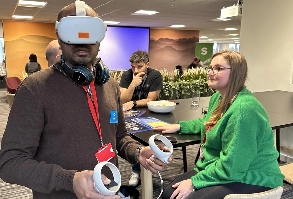 A person wearing a VR headset and holding controllers stands in an office space, highlighting the versatility of virtual tools, while three people sit and chat at a table in the background. Headphones hang around the VR users neck.