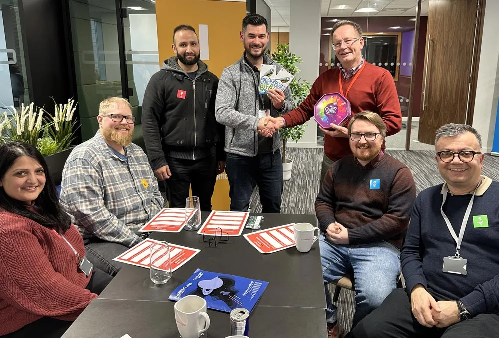 Seven people are gathered around a table in an office, celebrating collaboration. Two men shake hands—one with a trophy, the other with a box of chocolates—while others smile. Papers, mugs, and a can are scattered on the table.