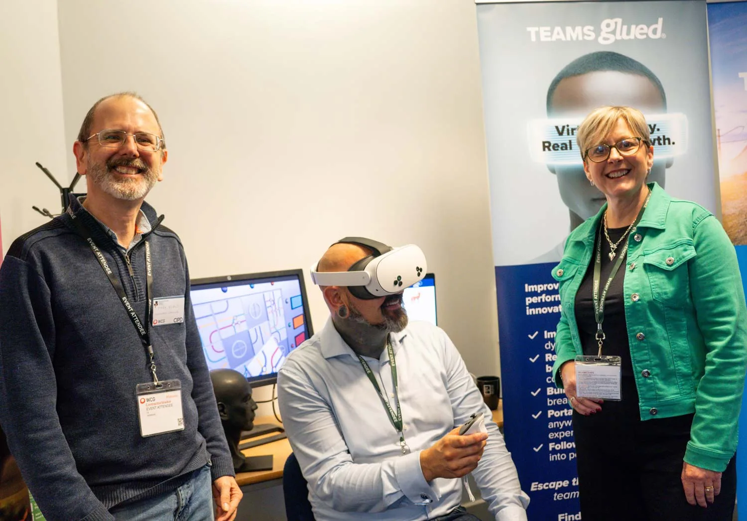 Three adults at a tech event engage in VR team building; one sits with a VR headset and controller while two stand smiling beside him. A computer and Teams Glued branding banner are visible in the background.