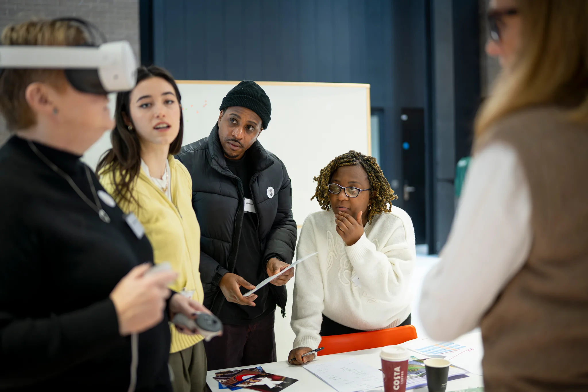 Four people gather around a table with papers and coffee cups. One person wears a VR headset and holds a controller, whilst the others watch, discussing insights into behaviour under pressure in a modern indoor setting.