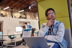 A man wearing glasses and a denim jacket sits with a laptop on his lap, thoughtfully looking to the side in a modern open-plan office—perhaps pondering why your B2B website isn’t converting and considering brand fixes that work.