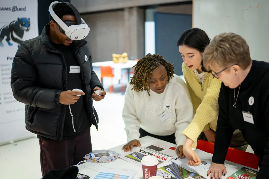 Four people work together at a table covered with papers and charts during a Teams Glued team building event. One person wears a virtual reality headset, while the others discuss documents, appearing focused and engaged in teamwork.