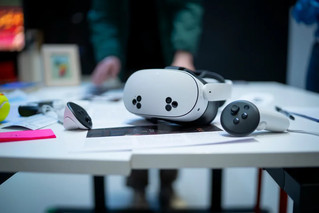 A virtual reality headset and two controllers rest on a white table covered with papers, suggesting preparations for a Teams Glued team building event, whilst a blurred person stands in the background.