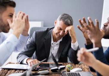 A group of five people sit around a table in a meeting room. Four are actively talking and gesturing, while one man in the center looks frustrated and rests his head on his hand—highlighting workplace conflict even in high-performing teams. Laptops and papers are on the table.
