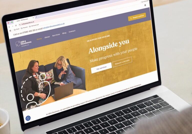 A person uses a laptop displaying a business website with a gold banner that reads “Alongside you: Make progress with your people.” The homepage highlights brand creation for HR business as two women on a couch talk and take notes.