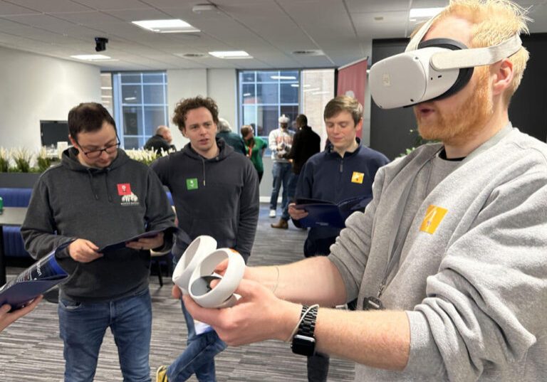 Four men stand in an office; one wears a VR headset and holds controllers while others watch him and look at Glued team building booklets. Casually dressed, they are engaged in a technology demonstration.