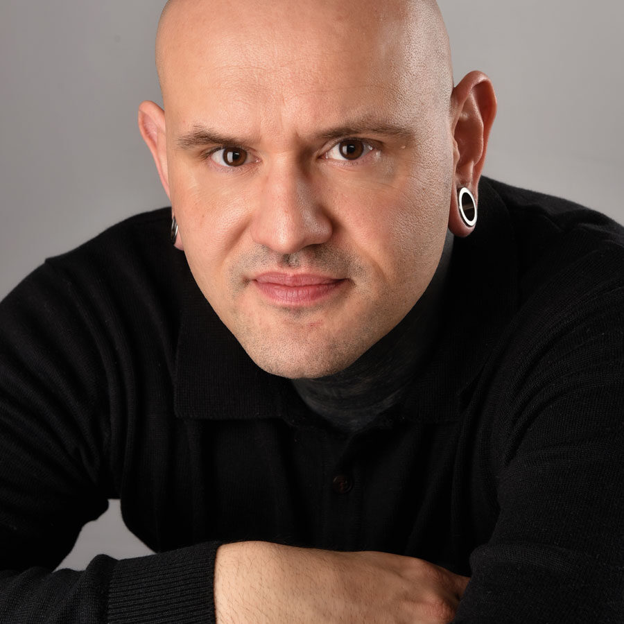 dw_small A bald person with gauged earrings, wearing a black shirt, poses confidently, arms crossed on a table against a neutral gray background. The calm and focused expression mirrors the poised professionalism of Glued Limited, a marketing agency in Warwickshire.