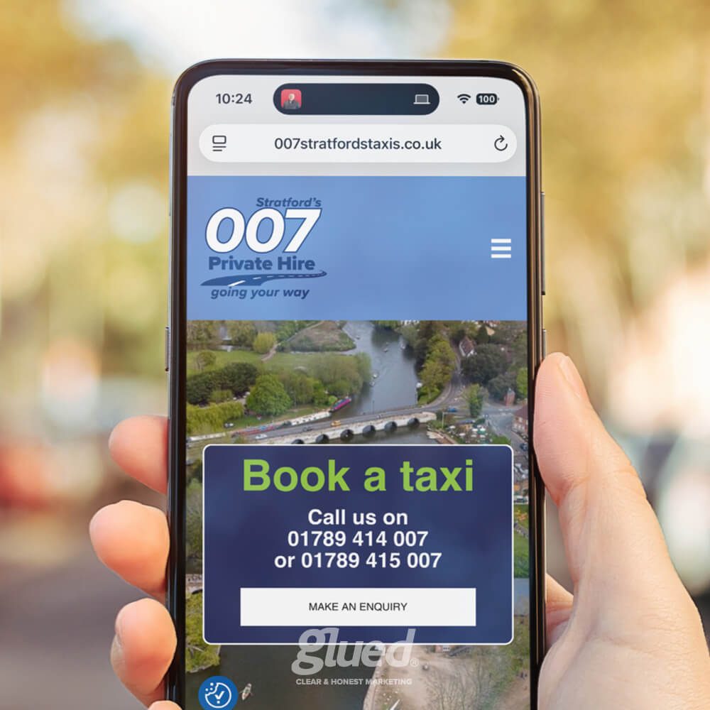 Glued Limited creative work: A hand holding a smartphone displaying the 007 Stratford Private Hire taxi service website, showing options to book a taxi and contact phone numbers, with a blurred outdoor background.