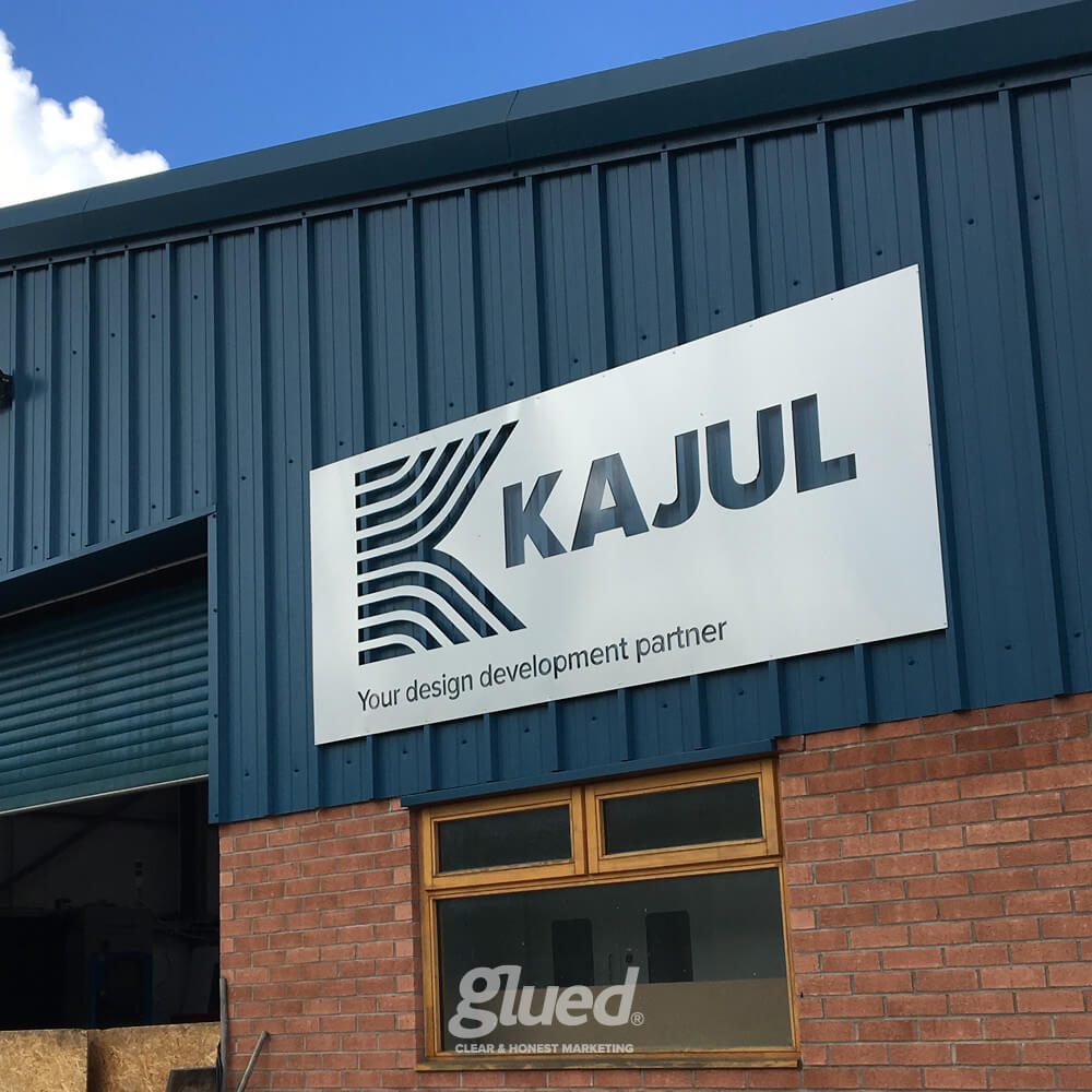 Glued Limited creative work: A large sign on a blue and red brick building reads KAJUL Your design development partner with a stylised logo. Below is a glass door with glued. CLEAR & HONEST MARKETING printed on it.