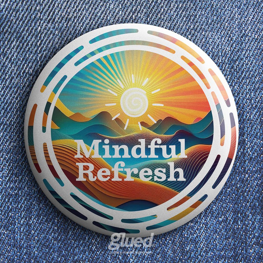 Glued Limited creative work: A round button on denim fabric features colourful abstract mountains, a radiant sun, and the words Mindful Refresh in bold letters. The bottom shows a faint glued logo.
