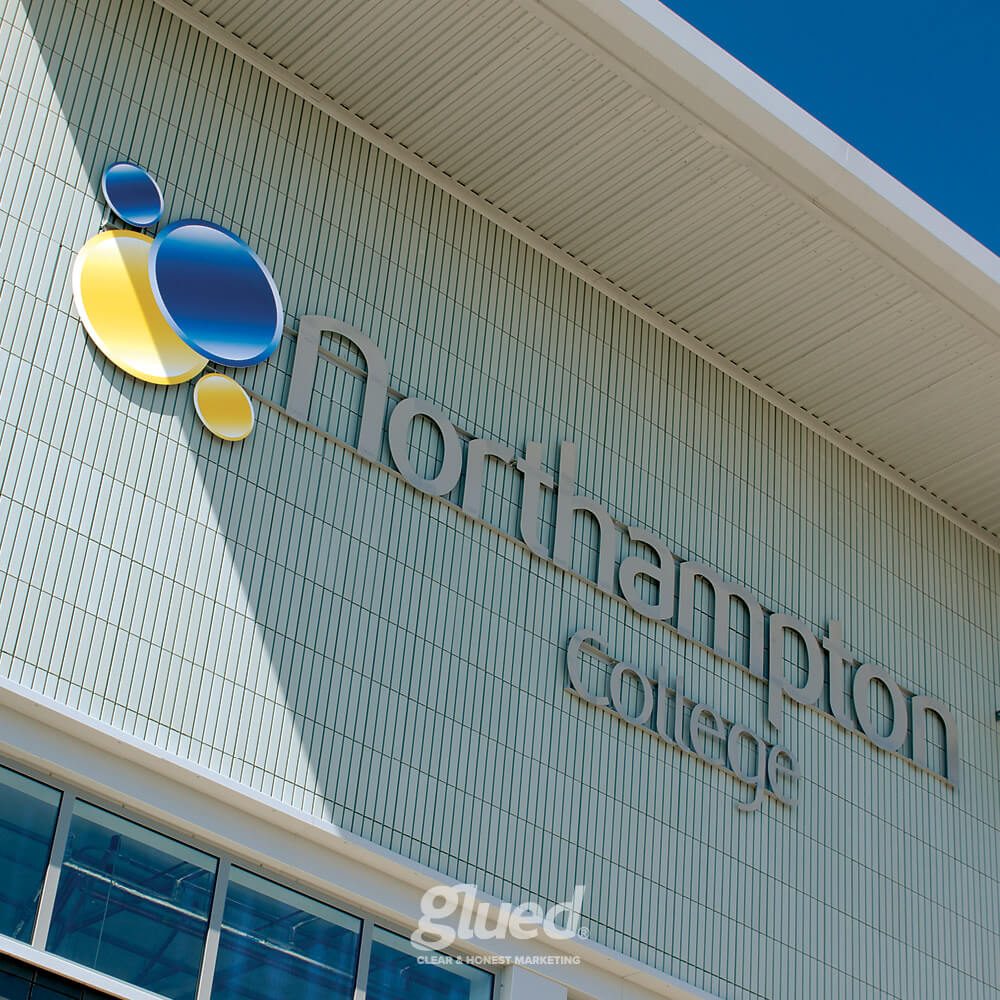 Glued Limited creative work: Exterior wall of Northampton College building with modern logo and name displayed in large letters; blue sky reflected in glass sections of the wall.