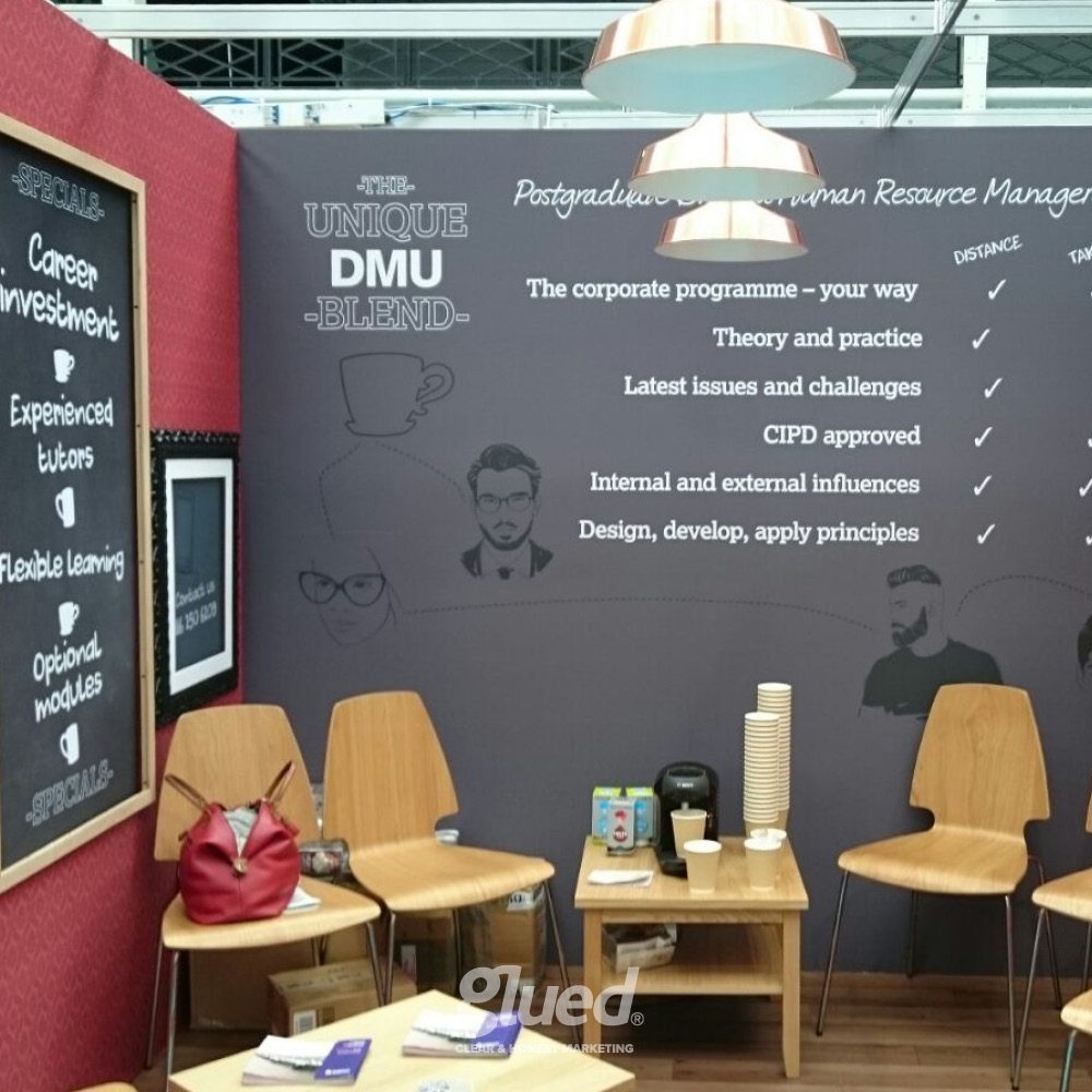 Glued Limited creative work: A cosy corner with chairs, a small table, books, bags, and a coffee cup sits before a decorated stand wall about human resource management programmes and educational benefits—perfect for marketing campaigns to reach like-minded people.