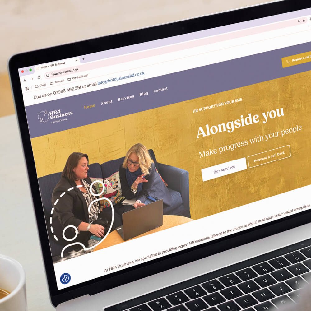 Glued Limited creative work: A laptop screen displays the homepage of an HR consulting WordPress website design and build at affordable prices. Two women sit on a sofa, discussing something whilst looking at the laptop. The website headline reads, “Alongside you. Make progress with your people.”.
