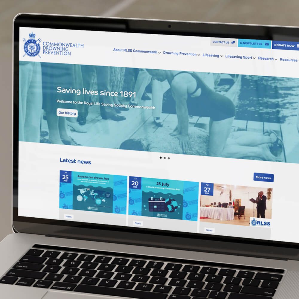 Glued Limited creative work: A laptop screen displays the Commonwealth Drowning Prevention WordPress website design and build at affordable prices, featuring a banner with historical lifeguard photos, navigation menus, and a news section with event updates.